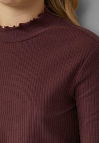 Nahaufnahme einer Person in einem gerippten, dunkelvioletten Langarmoberteil mit einem gerafften Stehkragen, deren blonde Haare auf der Schulter sichtbar sind.