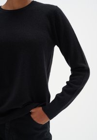 Schwarzer Strickpullover mit rundem Halsausschnitt, langen Ärmeln und gerippten Bündchen. Weicher Stoff und figurbetonte Silhouette mit einer seitlichen Taschenoptik.
