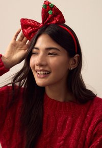 Rotes, mit Pailletten besetztes Stirnband mit großer Schleife, verziert mit grünen Stechpalmenblättern und Beeren. Kombiniert mit einem roten Pullover mit Zopfmuster.