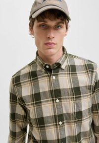 Camisa de estampado a cuadros en verde y marrón claro con cuello. La textura del tejido parece suave. Incluye un bolsillo en el pecho y botones blancos.