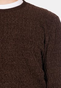 Primo piano di una persona che indossa un maglione lavorato a maglia di colore marrone scuro con texture sopra una maglietta bianca a girocollo.