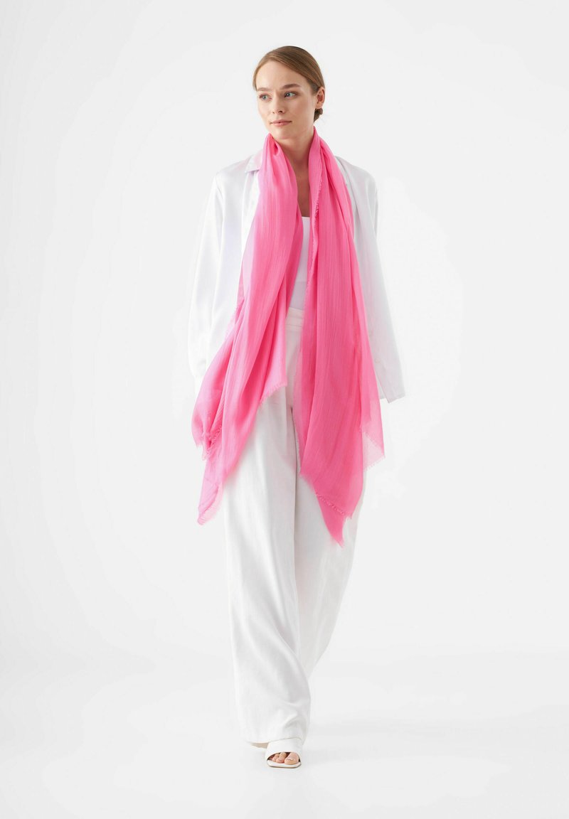 Écharpe rose légère avec une texture douce et transparente, drapée autour du cou, portée sur un haut blanc et un pantalon blanc à jambes larges.