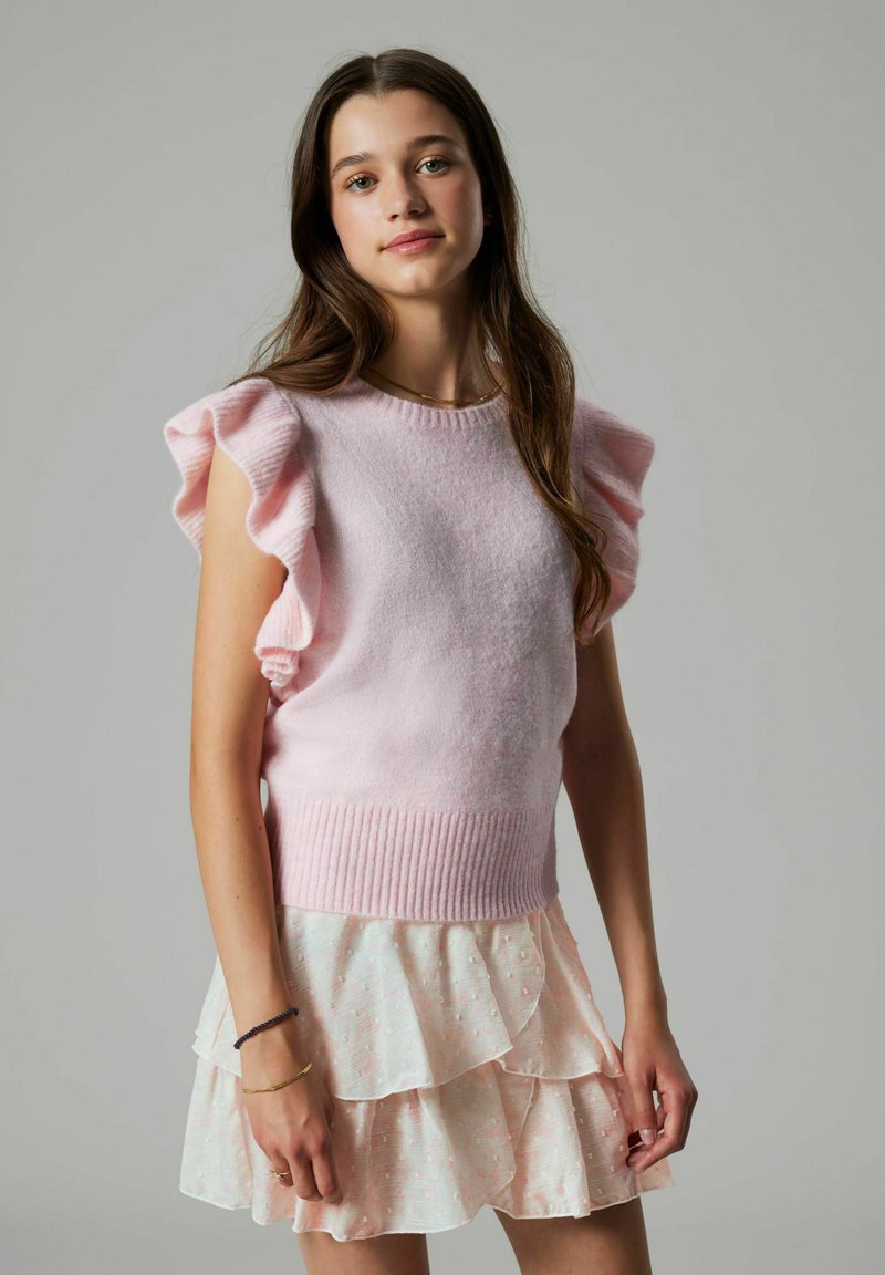 Jeune femme aux longs cheveux bruns portant un pull sans manches rose clair à volants et une jupe rose pâle à plusieurs couches, debout devant un fond gris uni.