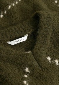 Tissu tricoté duveteux vert foncé avec de petits points blancs brodés et une étiquette de vêtement blanche indiquant NOELLA.
