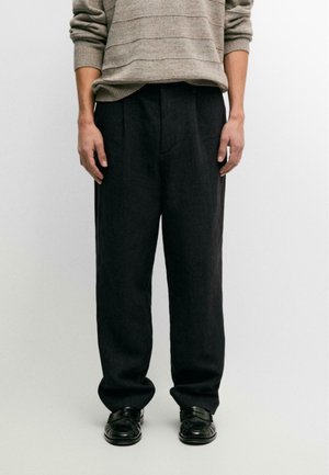 Mann trägt lockere schwarze Hose, schwarze Lederschuhe und einen beigen Pullover mit langen Ärmeln, dessen Ärmel hochgekrempelt sind, steht vor einem einfarbigen Hintergrund.