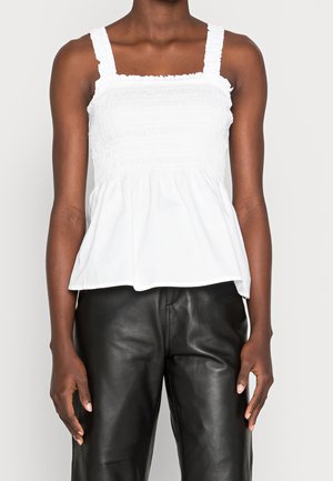 Vrouw die een witte mouwloze top met smockwerk en gerimpelde bandjes draagt, gecombineerd met zwarte leren broek, staand tegen een effen lichte achtergrond.