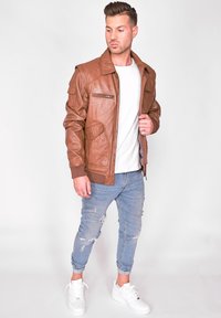 Brun læder bomberjakke med ribstrikkede manchetter, lynlåslommer og klassisk krave, parret med lyseblå slidte jeans og hvide sneakers.