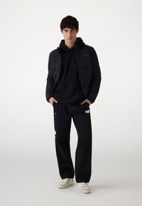 Sort dunjakke med lynlås, hætte-svart sweatshirt og løse sorte bukser. Accenter inkluderer logodetaljer og hvide sneakers.