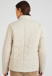 Giacca trapuntata beige con un sottile motivo a chevron, dotata di colletto alto e una texture morbida e liscia. Senza hardware o accenti visibili.