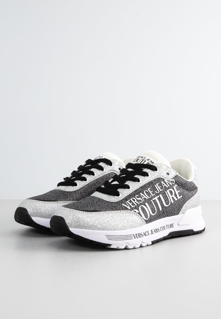 Zapatos deportivos en negro y gris con parte superior de malla, acentos metálicos y marca blanca. Presentan cordones negros y una suela texturizada.