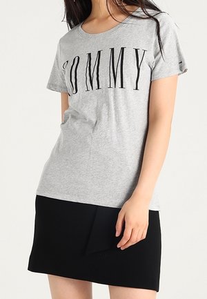 Grå kortærmet t-shirt med "TOMMY" trykt i store sorte bogstaver. Kombineret med en sort nederdel med en foldet akcentdetalje.