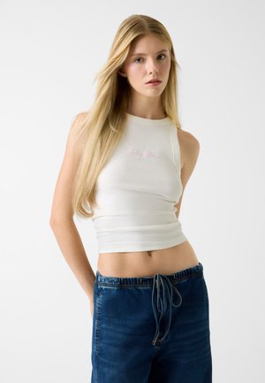 Witte geribbelde tanktop met een ronde halslijn en roze geborduurd detail, gecombineerd met blauwe jeans met een aantrekkoord en een losse pasvorm.
