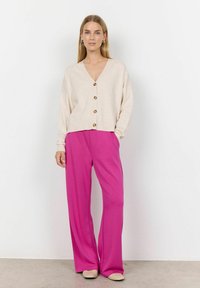 Beige button-up cardigan with a V-neck and long sleeves, paired with high-waisted, wide-leg pink trousers and light-colored flat shoes.