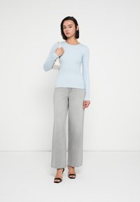 Svetlo modra majica z dolgimi rokavi, sive široke kavbojke, črne sandale z visokimi petami, drži majhno belo torbico na enobarvnem ozadju.