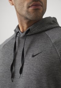 Gros plan d’un homme portant un sweat à capuche gris Nike avec des cordons noirs et un logo Nike visible sur la poitrine.