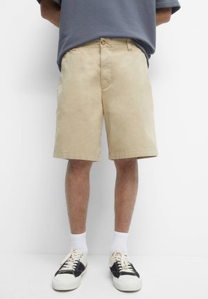 Shorts beiges en coton, au design classique avec une fermeture à bouton, des poches latérales et un ourlet droit. Portés avec des chaussettes blanches et des baskets noires.