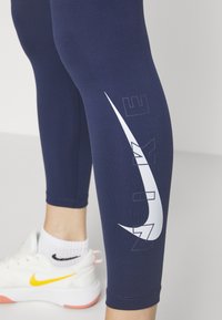 Granatowe legginsy z białym logo Nike w kształcie swooshu i napisem "NIKE". Miękki, elastyczny materiał o długości do połowy łydki i z mankietami przy kostkach. Białe sneakersy z żółtym akcentem.