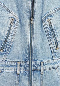 Veste en jean bleu clair à l'avant avec une fermeture éclair centrale, deux poches poitrine zippées et des passants de ceinture cousus à la taille.