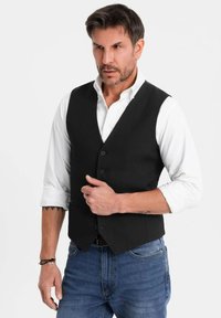 Uomo con capelli corti e scuri che indossa una camicia bianca arrotolata, un gilet nero con bottoni, jeans blu e bracciali, in piedi di fronte a uno sfondo chiaro e semplice.