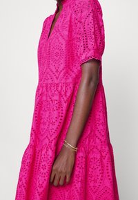 Roze geborduurde jurk met een V-hals, korte pofmouwen, een gelaagde rok en ingewikkelde oogjespatronen. Gouden armband zichtbaar om de pols.