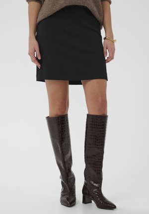 Ženska, oblečena v črno mini krilo, rjave teksturirane škornje do kolen, udoben rjavi pulover in zlata zapestnica na desni zapestju.