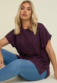 Kortärmad, djuplila topp med texturerad, ribbad tyg. Löst sittande med sidoklyftor, parat med ljusblå jeans.