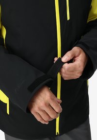 Schwarz-gelbe Jacke mit einem Reißverschluss vorne, verstellbaren Klettverschlussärmeln und matter Oberfläche. Sichtbare Nähte betonen das Design.