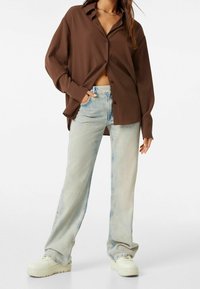 Camicia marrone a maniche lunghe con bottoni frontali, indossata sopra jeans a gamba larga blu chiaro, abbinata a sneakers bianche.