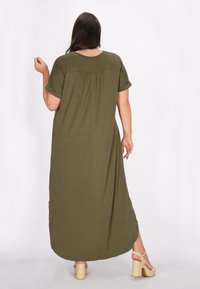 Rochie de culoare verde măslin, până la glezne, cu mâneci scurte răsucite, detaliu cu strângere la spate și tiv rotunjit. Asociată cu sandale cu platformă bej, masive.