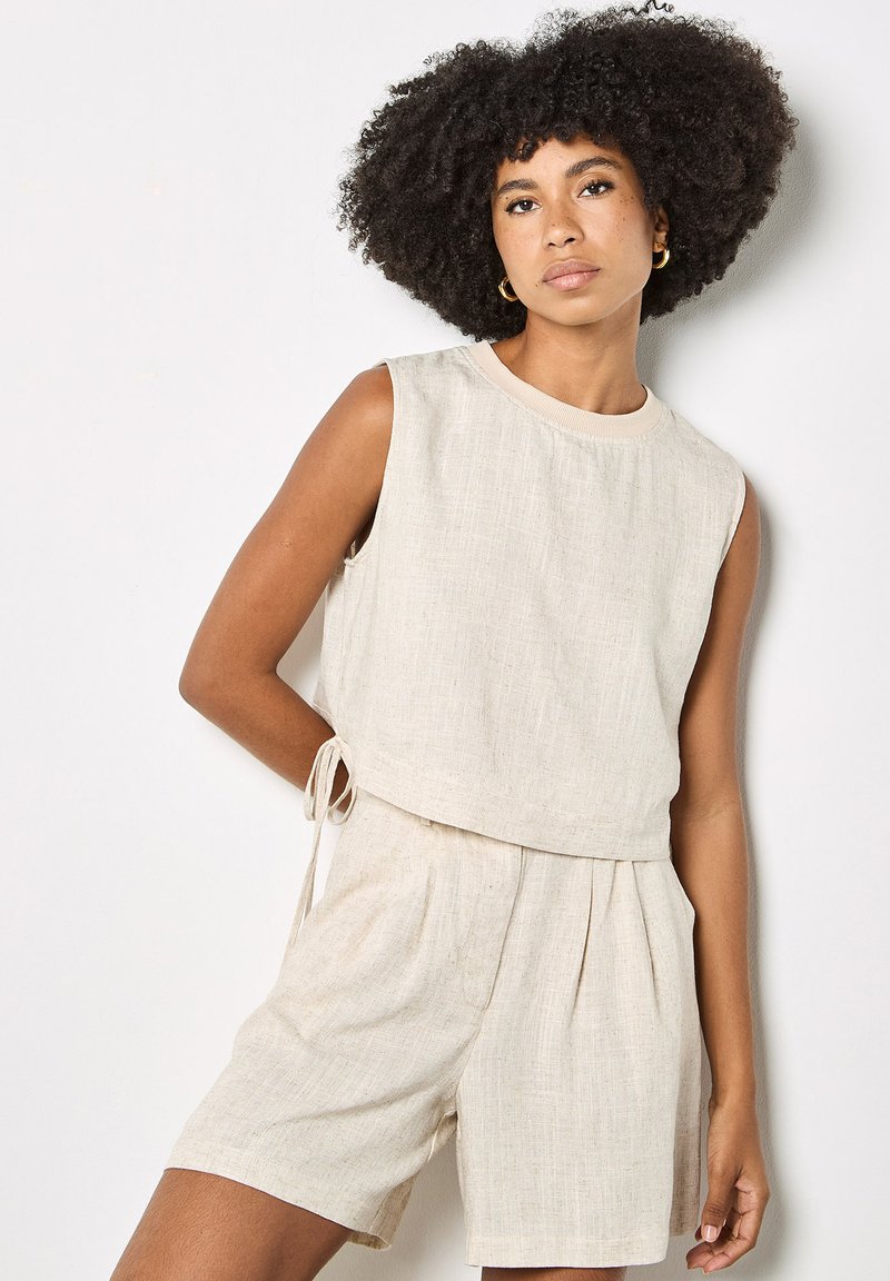 Femme aux cheveux bouclés volumineux portant un haut sans manches en lin beige et un short assorti, posant avec la main derrière le dos contre un mur blanc.
