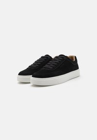 Black suede sneakers with a low profile, white rubber sole, rounded toe, and black laces. Features a tan leather accent inside.