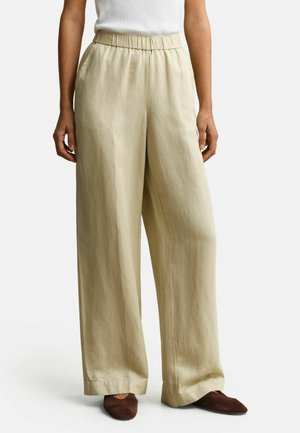 Person trägt lose beigefarbene Hose mit weitem Bein und elastischem Bund sowie braune flache Schuhe, steht vor einem einfarbigen Hintergrund.