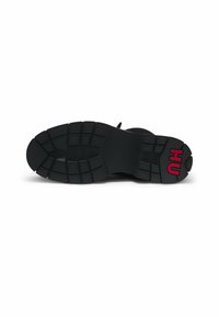 Semelle de botte noire avec des rainures profondes, arborant un logo rouge "HU" près du talon, photographiée sur un fond blanc.
