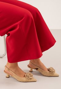 Zapatos slingback beige con textura trenzada, punta afilada y un acento floral. Combinados con pantalones rojos de pierna ancha, mostrando un primer plano del calzado.