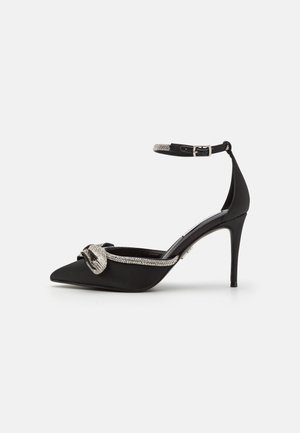 Chaussure noire à talon haut pointu avec bride à la cheville et ornement nœud en strass argenté sur l'avant.