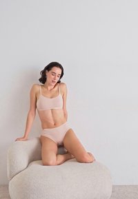Lichtroze naadloze bralette en bijpassende bikinibroekjes, zachte stof, minimalistisch ontwerp. Model zit op een gebogen, gestructureerde beige stoel.