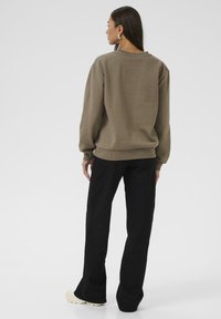 Hanorac oversized maro cu manșete și tiv din material elastic, purtat împreună cu pantaloni negri evazați și adidași de o culoare deschisă. Textură a materialului neted.