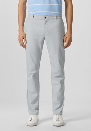 Homme portant un pantalon slim gris clair, des baskets blanches et une chemise rayée bleu et blanc, debout devant un fond uni.