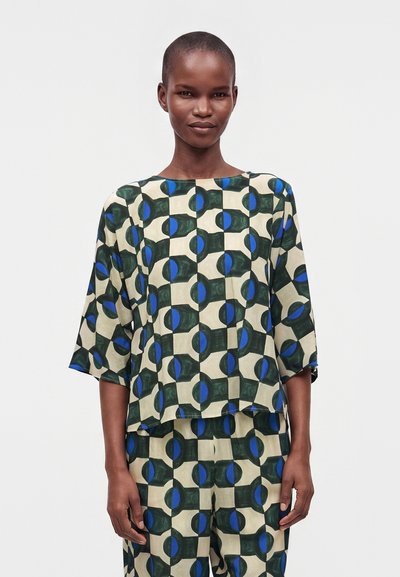 Femme portant un haut et un pantalon assortis avec un motif géométrique de cercles verts, bleus et beiges, debout devant un fond uni.