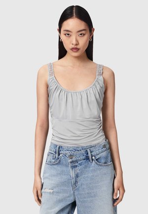 Jonge vrouw met lang donker haar, gekleed in een lichtgrijze getailleerde tanktop met kanten bandjes en een lichtblauwe, hooggetailleerde spijkerbroek.