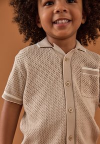 Beiges Strickshirt mit kurzen Ärmeln, vorne mit Knöpfen, einer Tasche und strukturierten Mustern. Einfacher Kragen mit kontrastierendem Besatz.