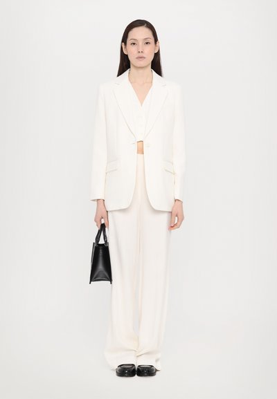Une femme se tient face à l'avant, portant un costume entièrement blanc avec un pantalon large et un blazer, tenant un petit sac à main noir dans sa main droite.