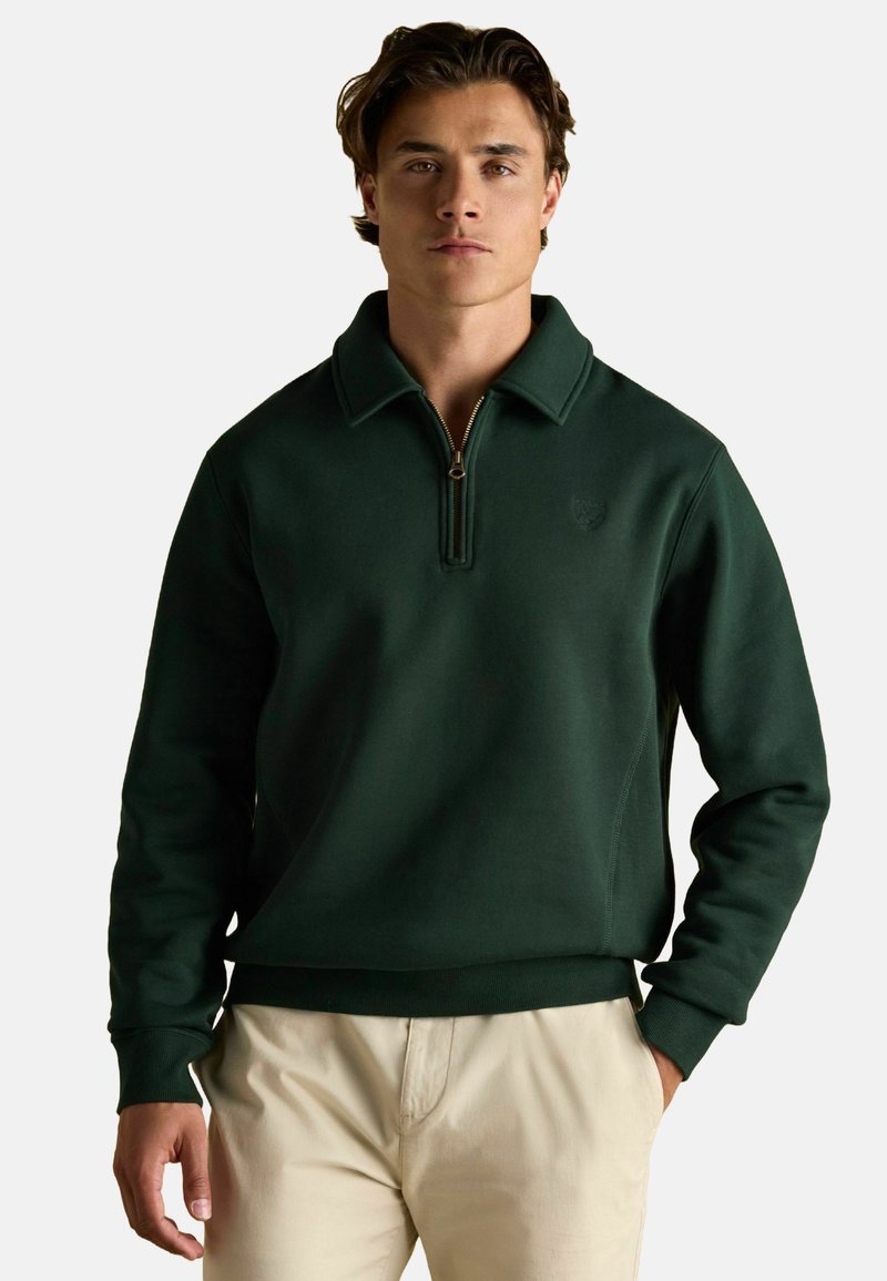 Zielony sweter z zamkiem przy kołnierzu, o teksturowanym wykończeniu i ściągaczach na mankietach, w zestawieniu z jasnymi spodniami.