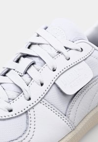 Baskets blanches avec une tige en cuir lisse, des panneaux en tissu texturé, des lacets plats et une semelle cousue. Comprend une étiquette logo bien visible.