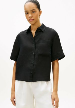 Mujer con camisa negra de manga corta con botones y un bolsillo en el pecho, y pantalones blancos de cintura alta, sobre un fondo liso.