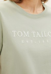 Lys grøn bomulds T-shirt med rund hals, der har broderet hvid tekst "TOM TAILOR" og "EST-1967" på forsiden.