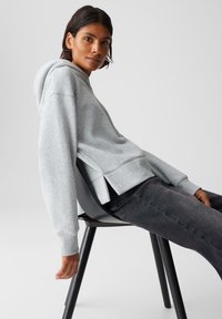 Persona appoggiata indietro su una sedia nera, indossa un felpa grigia chiara e jeans grigi scuri su uno sfondo neutro.