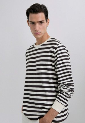 Camisa de manga larga a rayas en blanco y negro, fabricada con una tela suave. Presenta un escote redondo y puños acanalados, con un logo en la manga.