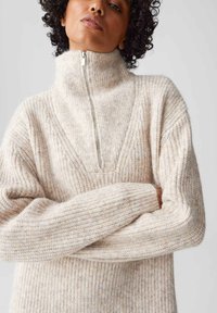 Maglione beige lavorato a maglia con collo a mezza zip, caratterizzato da una trama a costine e un design a forma di V. Tessuto morbido e caldo con una leggera variazione di colore.