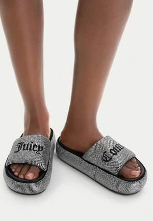 Piedi che indossano sandali slip-on argento scintillanti con "Juicy" scritto sulla fascia sinistra e "Couture" su quella destra.